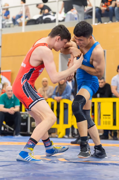 Two young wrestlers locked in a fierce clinch on the mat, red vs blue singlet, spectators watching in background.