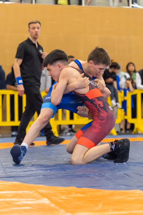 Two young wrestlers locked in a fierce takedown attempt on the mat, referee watching closely in the background.