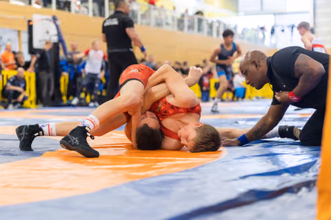 Two young wrestlers grapple on an orange mat as an intense coach leans in close, watching a pin attempt unfold.