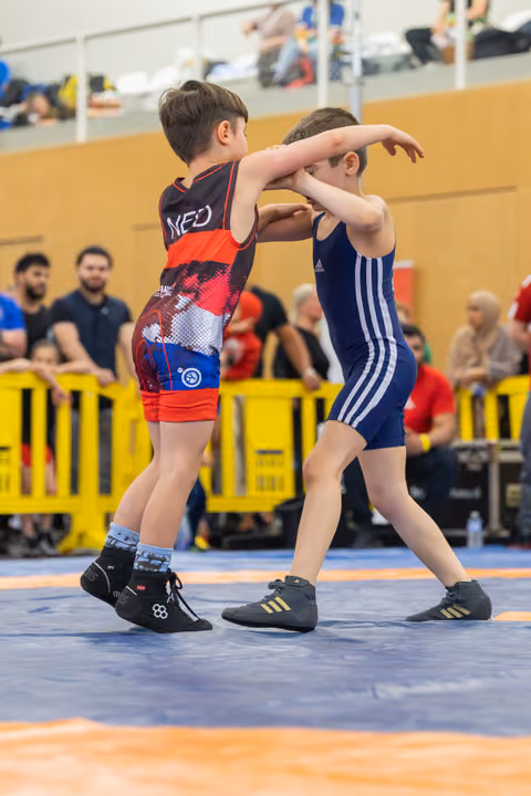 Two young boys locked in a wrestling hold on a blue mat, both focused and determined during an indoor bout.