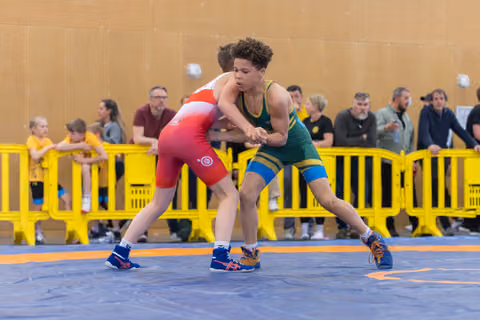 Two young wrestlers lock grips on the mat, the green-suited athlete driving forward with focused determination against his red-suited opponent.