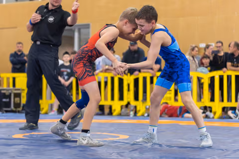 Two young wrestlers lock hands and press foreheads together in fierce concentration on a blue mat, referee watching.