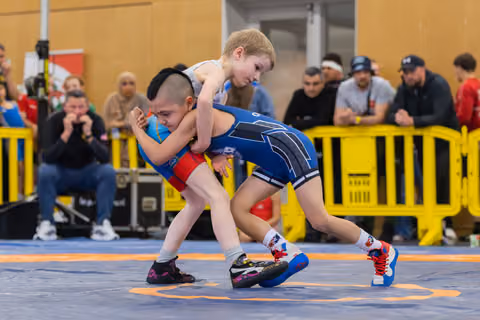Two young boys locked in an intense wrestling hold on a blue mat, surrounded by spectators at an indoor sports event.