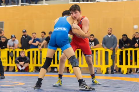 Teen wrestler in red singlet grips opponent in blue, both locked in intense standoff on competition mat.