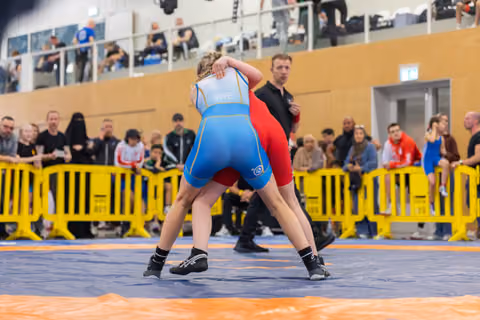 Swedish wrestler in blue singlet grapples opponent in red on mat, crowd watching from behind yellow barriers
