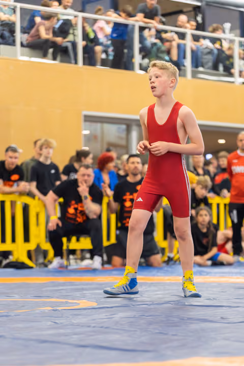 Young wrestler in red Adidas singlet stands focused on the blue mat, crowd watching from the stands behind.