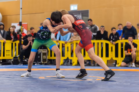 Two young wrestlers lock in a tight clinch on a blue mat, surrounded by spectators behind yellow barriers.