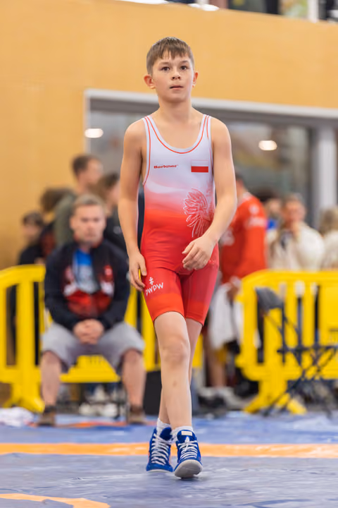 Focused young Polish wrestler walks confidently across the mat in red and white singlet, blue shoes, indoor arena.