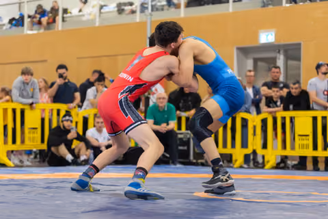 Two wrestlers locked in a fierce clinch on the mat, one in red, one in blue, crowds watching behind yellow barriers.
