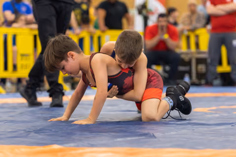 Two young boys wrestling intensely on a blue mat, one applying pressure from above while the other braces on all fours.