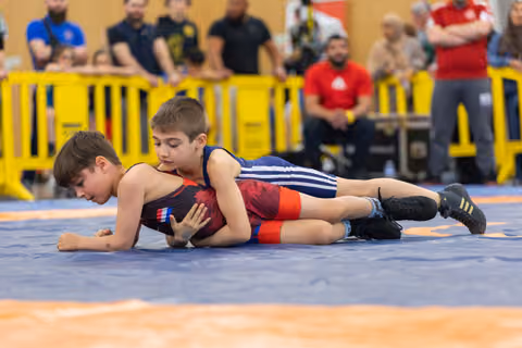 Young boy in blue singlet pins opponent to the mat, focused expression, amid a crowd of spectators at an indoor event.