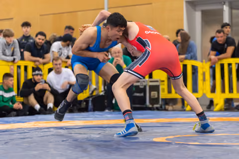 Two wrestlers clash on the mat, the blue-suited athlete lunging aggressively while the red-suited opponent braces firmly.