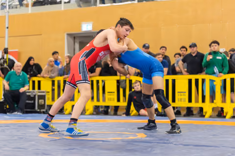 Two young wrestlers locked in an intense clinch on the blue mat, crowd watching behind yellow barriers.