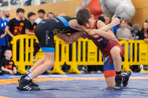 Two young wrestlers locked in a tight clinch, both leaning forward intensely during a youth match on a blue mat.