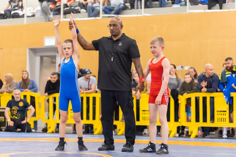 Referee raises the arm of a young boy in blue singlet declaring him winner, as defeated boy in red stands beside.