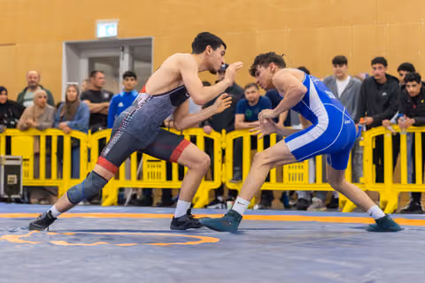 Two young wrestlers in a tense standoff on the mat, one in red-black, one in blue, crowd watching behind yellow barriers.