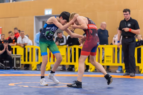 Two young wrestlers lock grips on a blue mat, both leaning in intensely, referee watching closely behind yellow barriers.