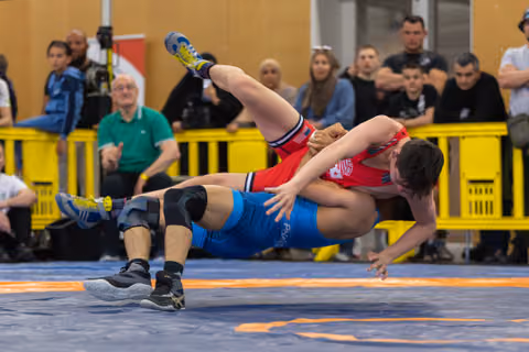 Wrestler in blue singlet executes a powerful throw, lifting red-clad opponent mid-air over the mat before spectators.