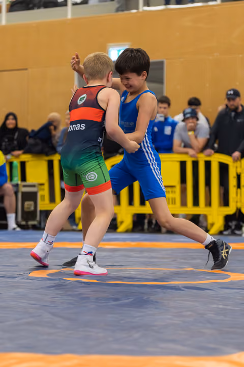 Two young boys locked in an intense wrestling bout on a blue mat, one in blue singlet lifting his leg aggressively.