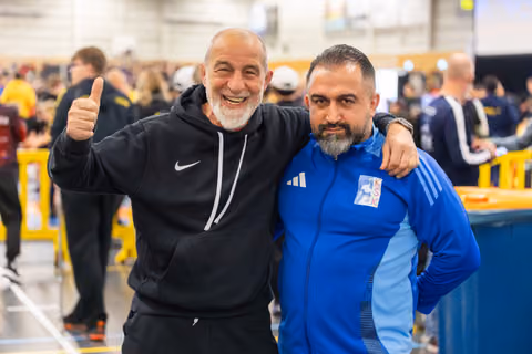Two coaches smile broadly together; the older man in a Nike hoodie gives a thumbs-up, arm around his colleague in a blue KSK jacket.