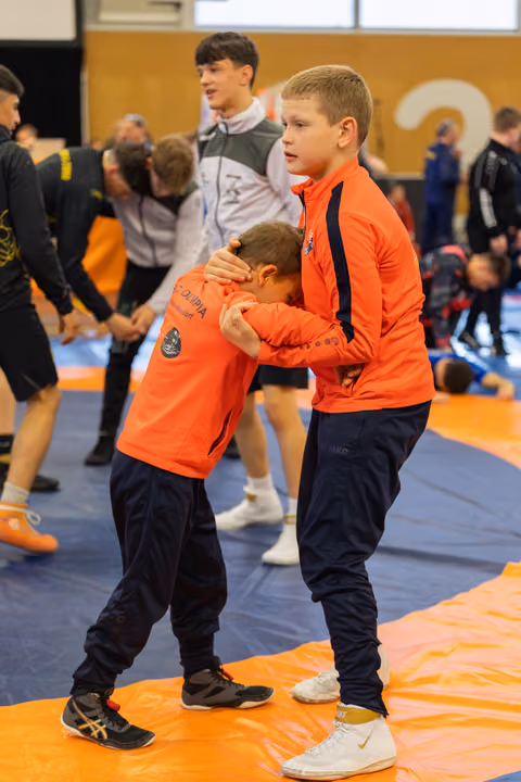 Two boys in orange jackets practice a wrestling hold on the mat, one bowing his head as the other guides firmly.