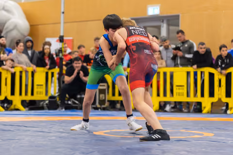 Two young wrestlers locked in a grapple on a blue mat, crowds watching behind yellow barriers at an indoor sports hall.