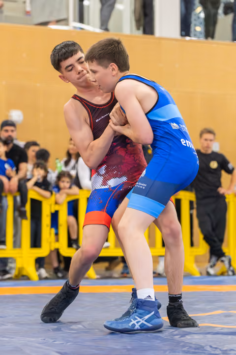 Two teenage wrestlers locked in a tight clinch, both straining with effort on the orange mat before a crowded gymnasium.