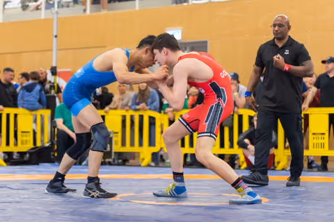 Two wrestlers in blue and red singlets lock grips on the mat, focused and tense, as a referee watches closely.