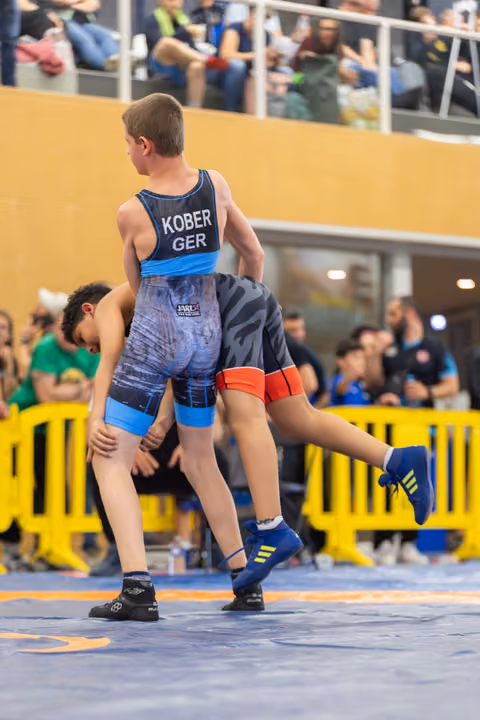 Young wrestler Kober (GER) lifts his opponent off the mat during an intense youth wrestling bout before a crowd.