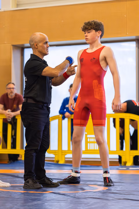 A bald coach points at a focused teenage wrestler in red singlet, offering pre-match guidance on the blue mat.