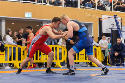Two wrestlers in red and blue singlets lock hands in an intense face-off on an orange mat, surrounded by spectators.