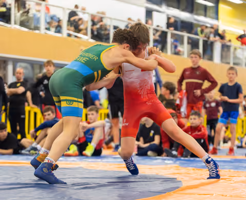 Two young wrestlers lock arms in fierce concentration, one in green-yellow, one in red, competing before a crowd.