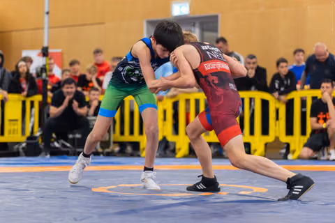 Two young wrestlers lock grips in intense clinch on blue mat, watched by crowd behind yellow barriers.