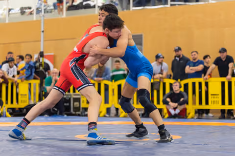 Two young wrestlers lock up in fierce competition, one in red, one in blue, straining forward on a blue mat.