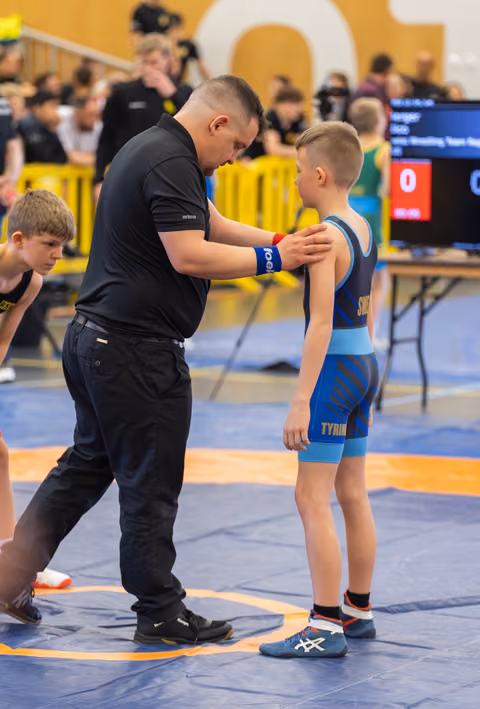 Coach places hand on young wrestler's shoulder, offering focused pre-match guidance on the blue mat during a youth event.