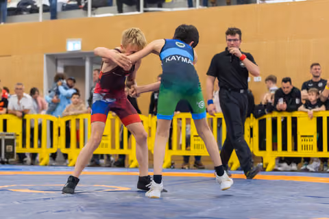 Two young boys in singlets grip each other intensely on a blue mat, referee watching closely behind yellow barriers.