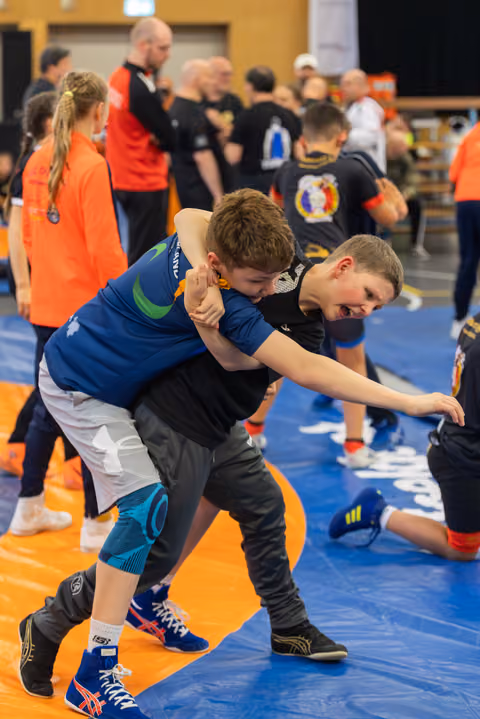 Two young boys wrestling intensely on a blue mat, one grimacing as he attempts a hold during a busy sports hall event.