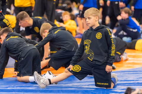 Blonde boy in Freiburg team gear stretches on the mat, looking composed while teammates warm up behind him.