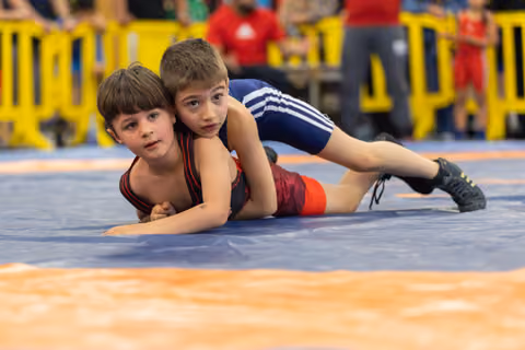 Two young boys wrestling on a blue mat, one in navy pinning the other in red, both showing intense concentration.