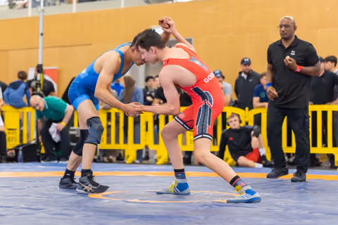 Two wrestlers grip hands in a tense standoff on the blue mat, referee watching closely from the sideline.