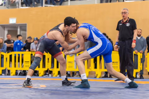 Two young wrestlers in focused stance, grappling on blue mat at indoor sports hall, referee watching nearby.