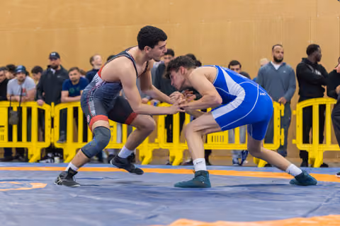 Two young wrestlers in black and blue singlets face off in a crouch, hands clasped, intense focus on the blue mat.