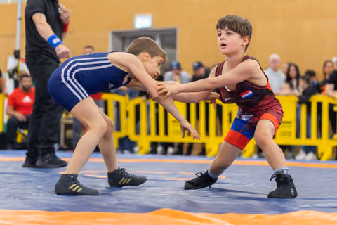 Two young wrestlers grip each other's wrists in a tense standoff on the mat, focused and determined.