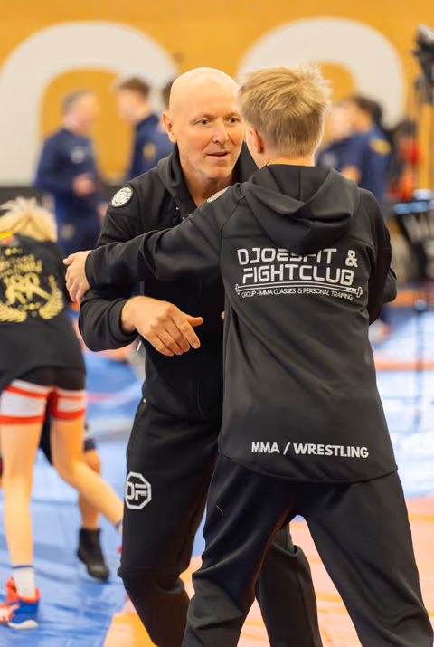 Bald coach embraces a young MMA wrestler in a Djoebfit Fight Club jacket, sharing a warm mentor moment on the mat.