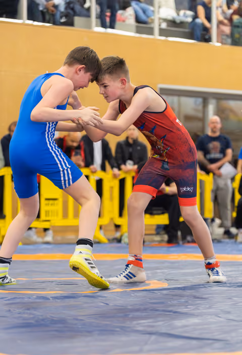Two young wrestlers grip each other's hands in a tense face-off on the mat, focused and determined.