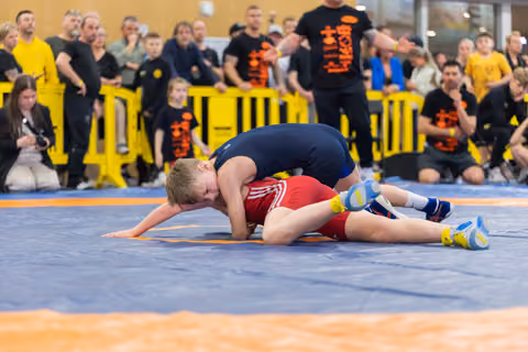 Young wrestler in red singlet being pinned by opponent in navy blue, crowd watching intently in background.