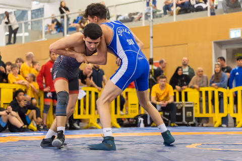Two young wrestlers lock grips in tense standoff on blue mat, crowd watching closely in packed indoor venue.
