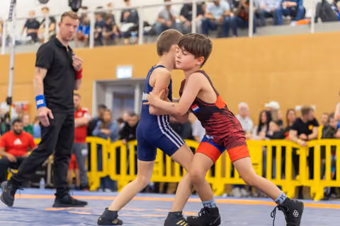 Two young boys wrestle intensely on the mat, locking grips, as a referee closely monitors nearby during a youth bout.
