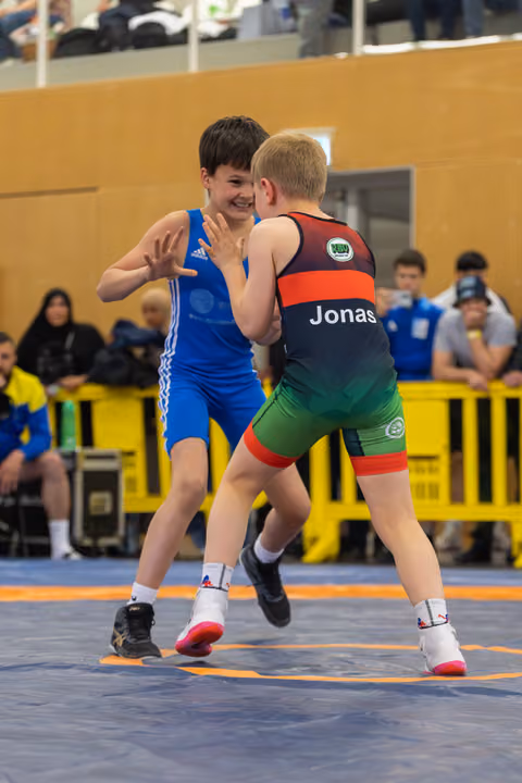 Smiling boy in blue Adidas singlet faces Jonas in black-red-green singlet on blue mat, indoor gym.