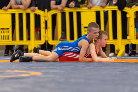Two boys wrestle on a gray mat, one in blue controlling the other in red, both showing fierce concentration.
